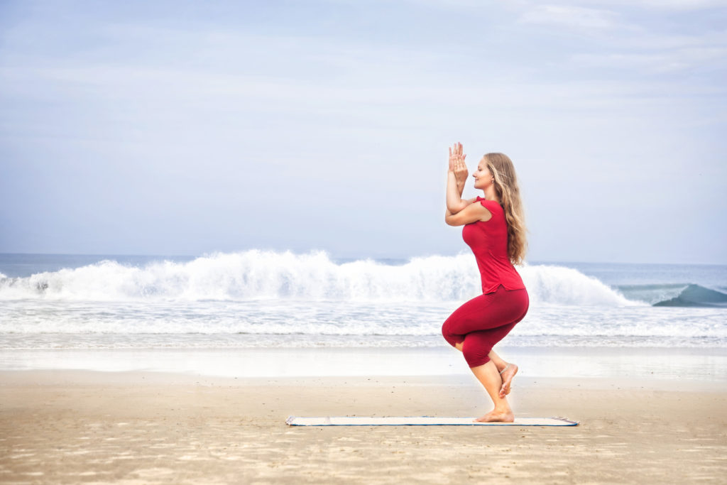 Yoga Garudasana eagle pose by young woman with long hair in red cloth on the beach at ocean background